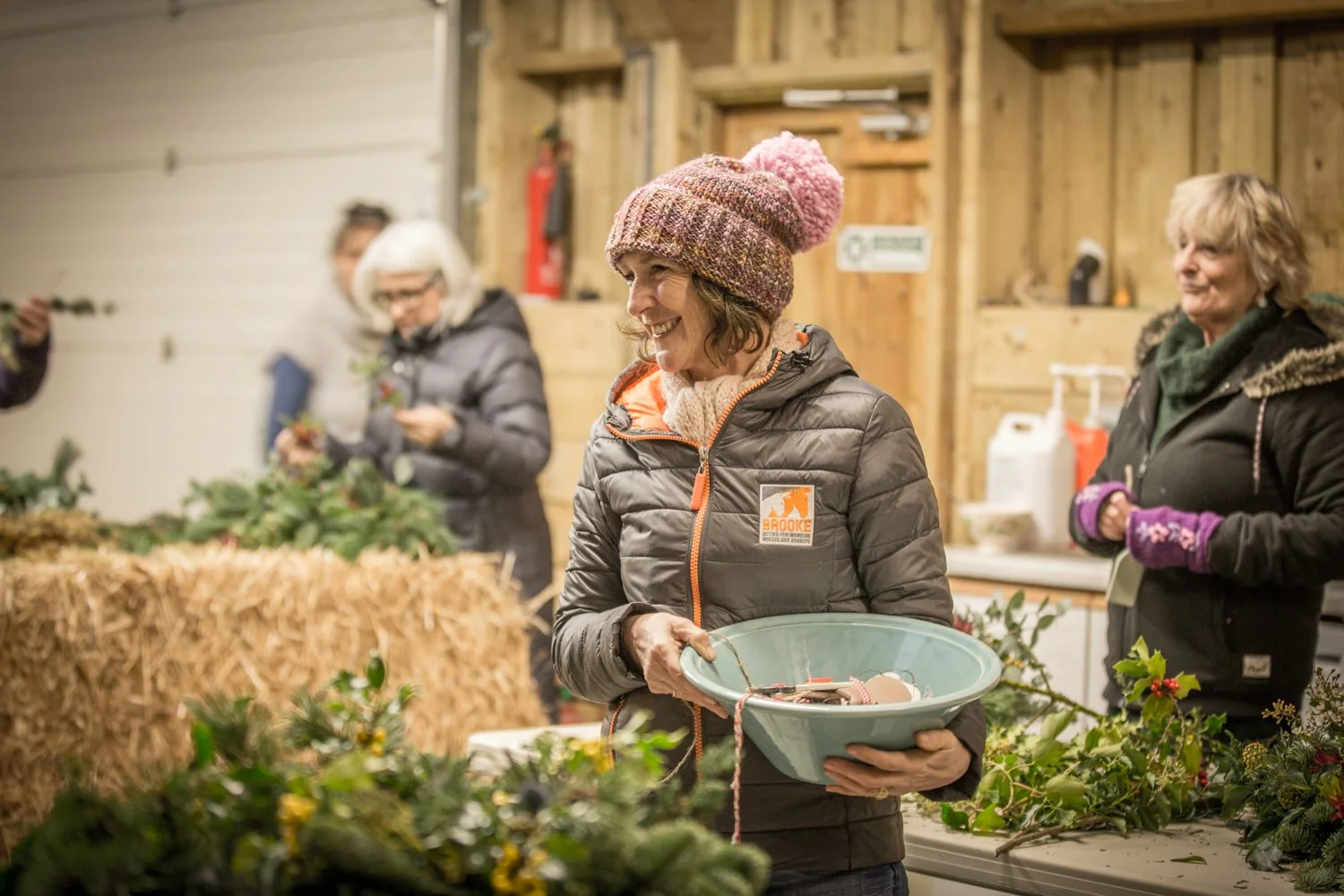 A woman in a pink knit hat and gray puffer jacket holding a green bowl, smiling inside a rustic wooden workshop decorated with holiday greenery and hay bales, with other women in winter clothing in the background.