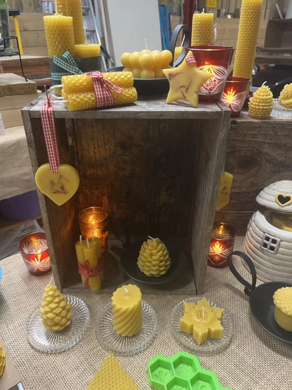 Decorative display of yellow candles, beeswax, and candle-shaped ornaments on a rustic wooden table, with candles in glass holders, a star-shaped wax decoration, and honeycomb or beehive-shaped candles, illuminated by candlelight.