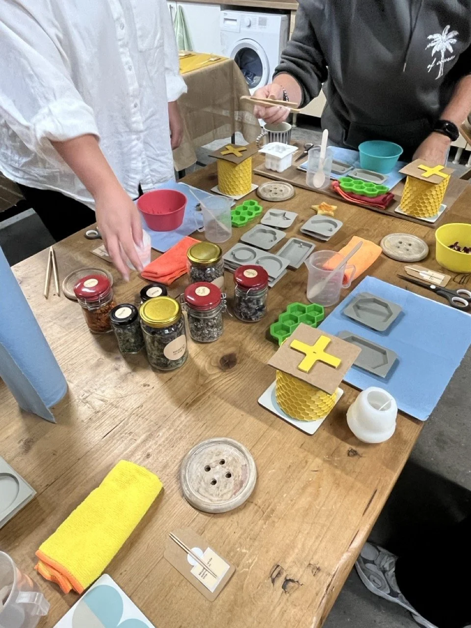 Table with colorful craft supplies including jars of spices, silicone molds, paper honeycomb holder with yellow and gray pieces, scissors, cloth, and various small tools in a room with washing machine in background.