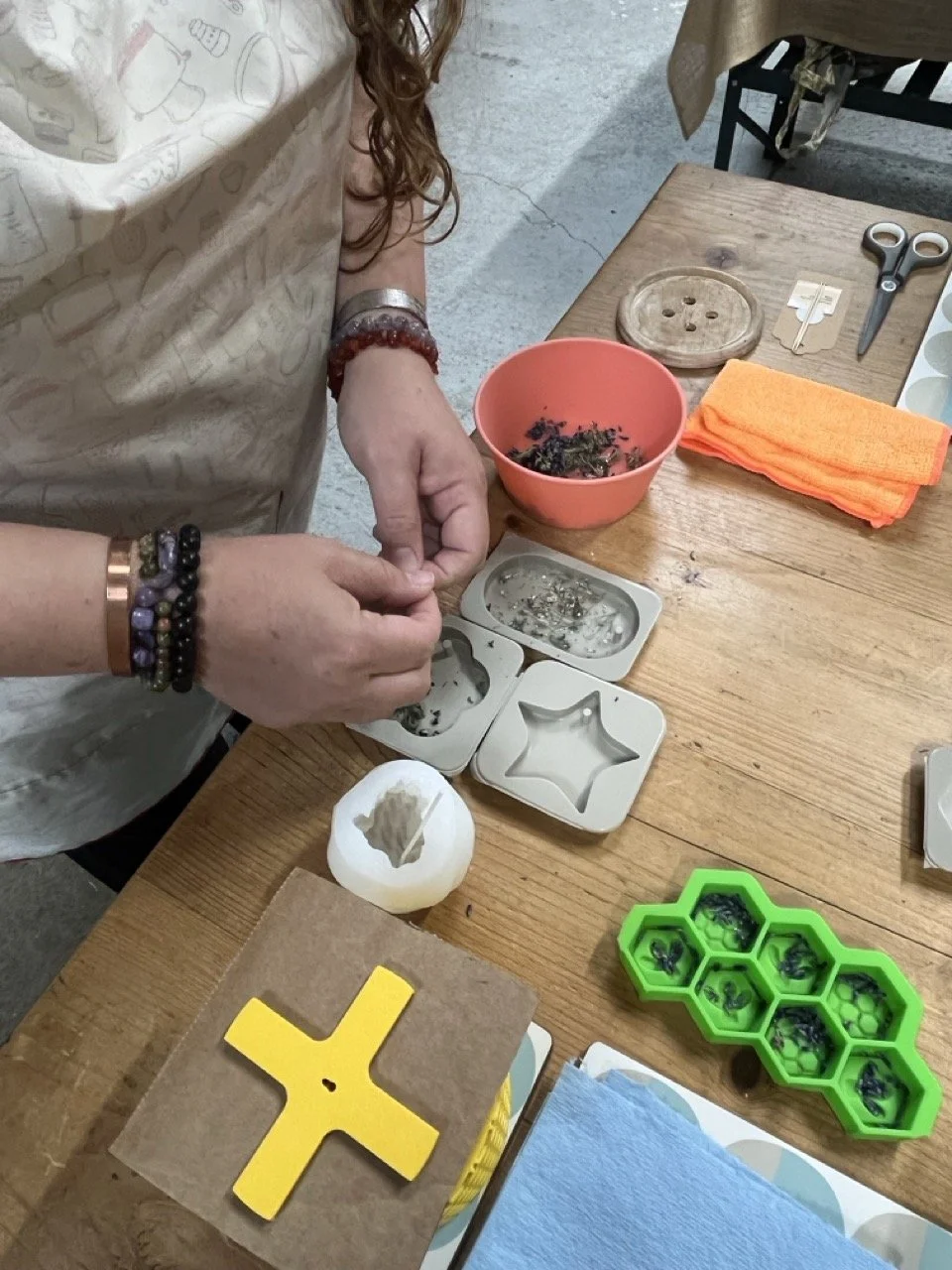 Person making candles on a wooden table, surrounded by crafting tools, a bowl with dried herbs, scissors, a candle, and small objects.