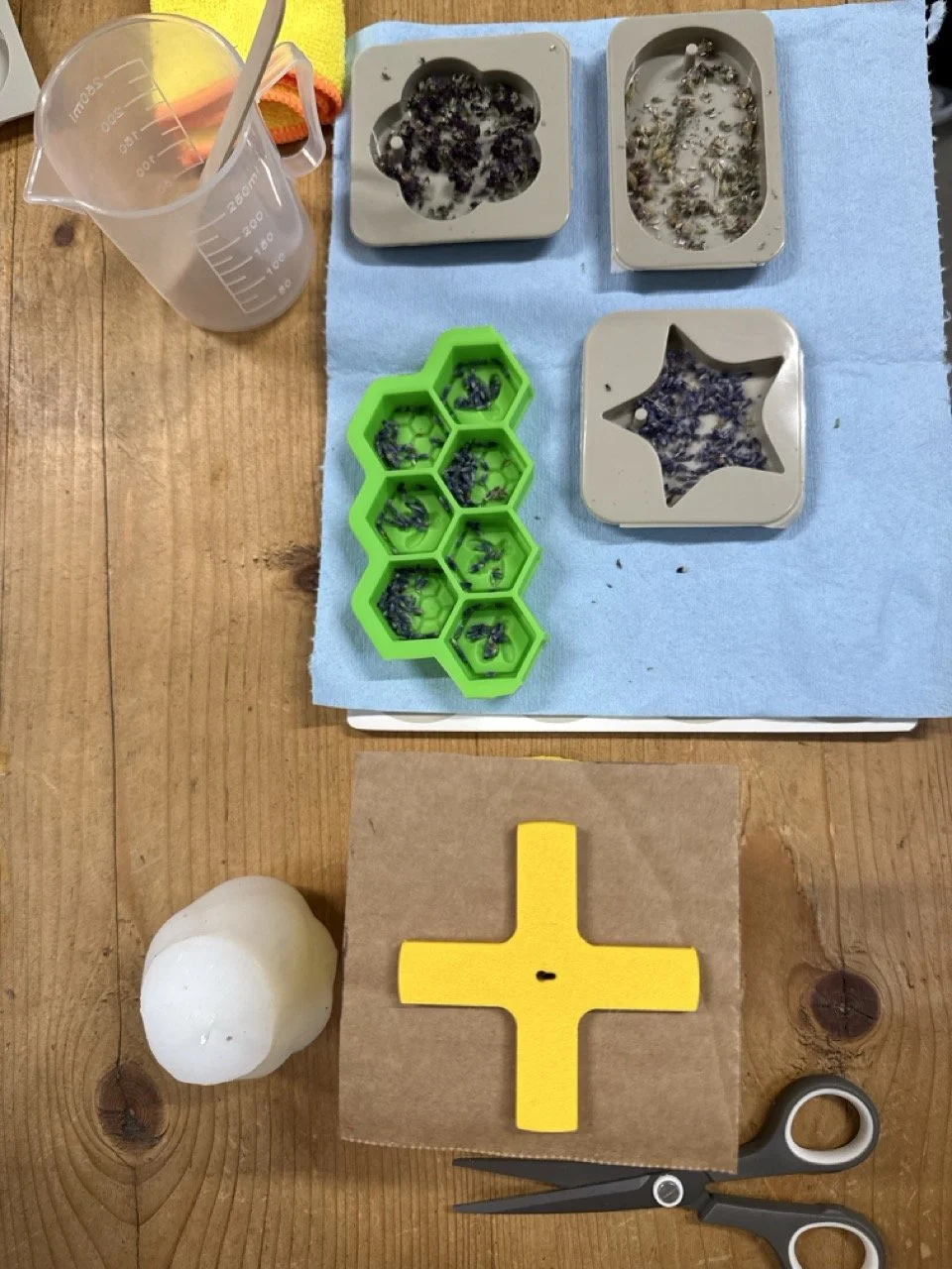 Lavender soap, a green hexagon-shaped soap mold filled with lavender soap, a clear measuring cup with a straw, a yellow cross-shaped soap mold, a pair of scissors, a white soap, and a blue paper towel on a wooden table.