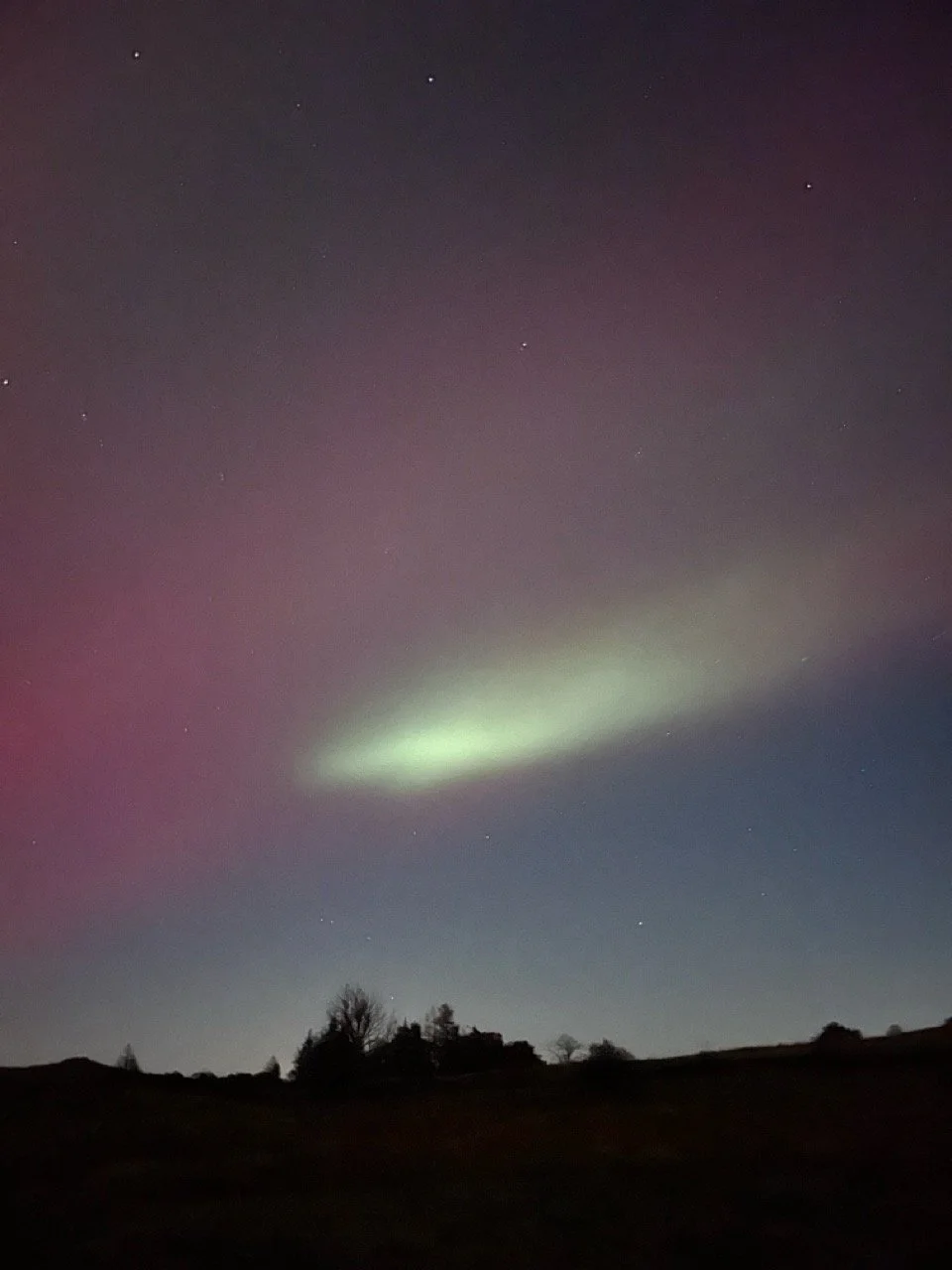 Night sky with visible northern lights and stars, silhouette of trees and hillside in the foreground.