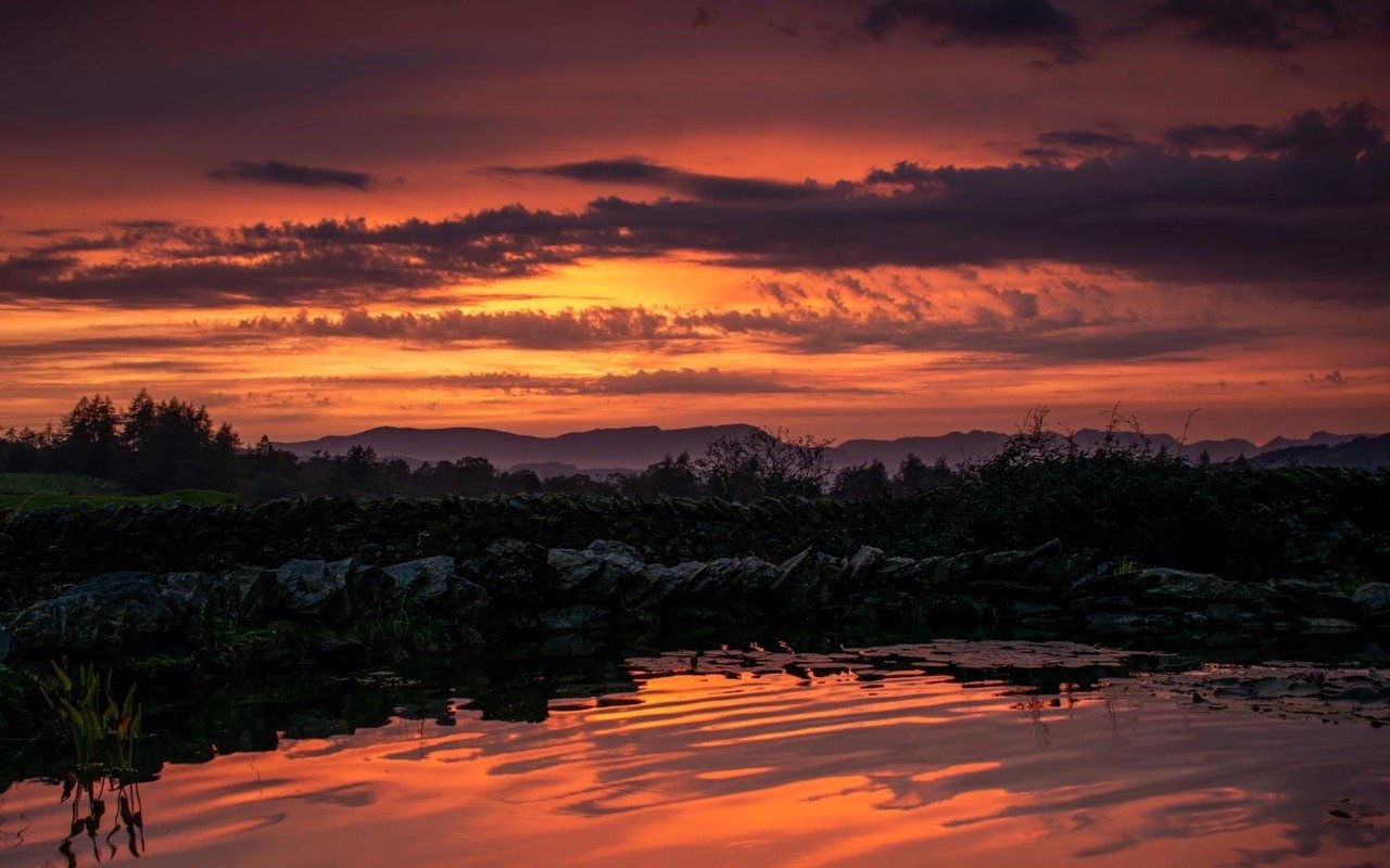 Sunset over a landscape with a rocky shoreline, calm water reflecting the colorful sky, and distant hills and trees.