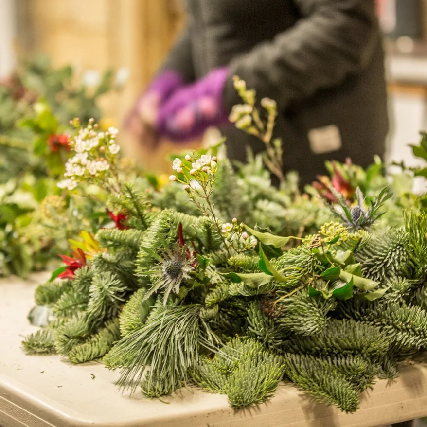 A person working on floral arrangement with pine branches, holly, and other holiday greenery at a table.