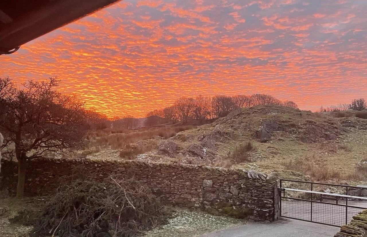 A sunset sky with vibrant orange and pink clouds over a grassy, rocky hillside, with a stone wall and a metal gate in the foreground.