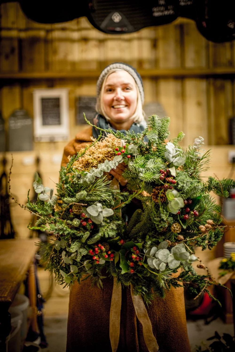 Woman holding a Christmas wreath made of various green foliage, berries, and pinecones in a wood-paneled workshop.