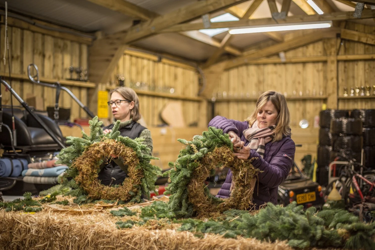 Two women decorating Christmas wreaths with evergreen branches in a rustic wooden workshop.