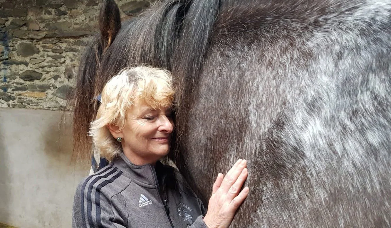 A woman with blonde hair gently touching a horse's side with her eyes closed, in a stable with stone walls.