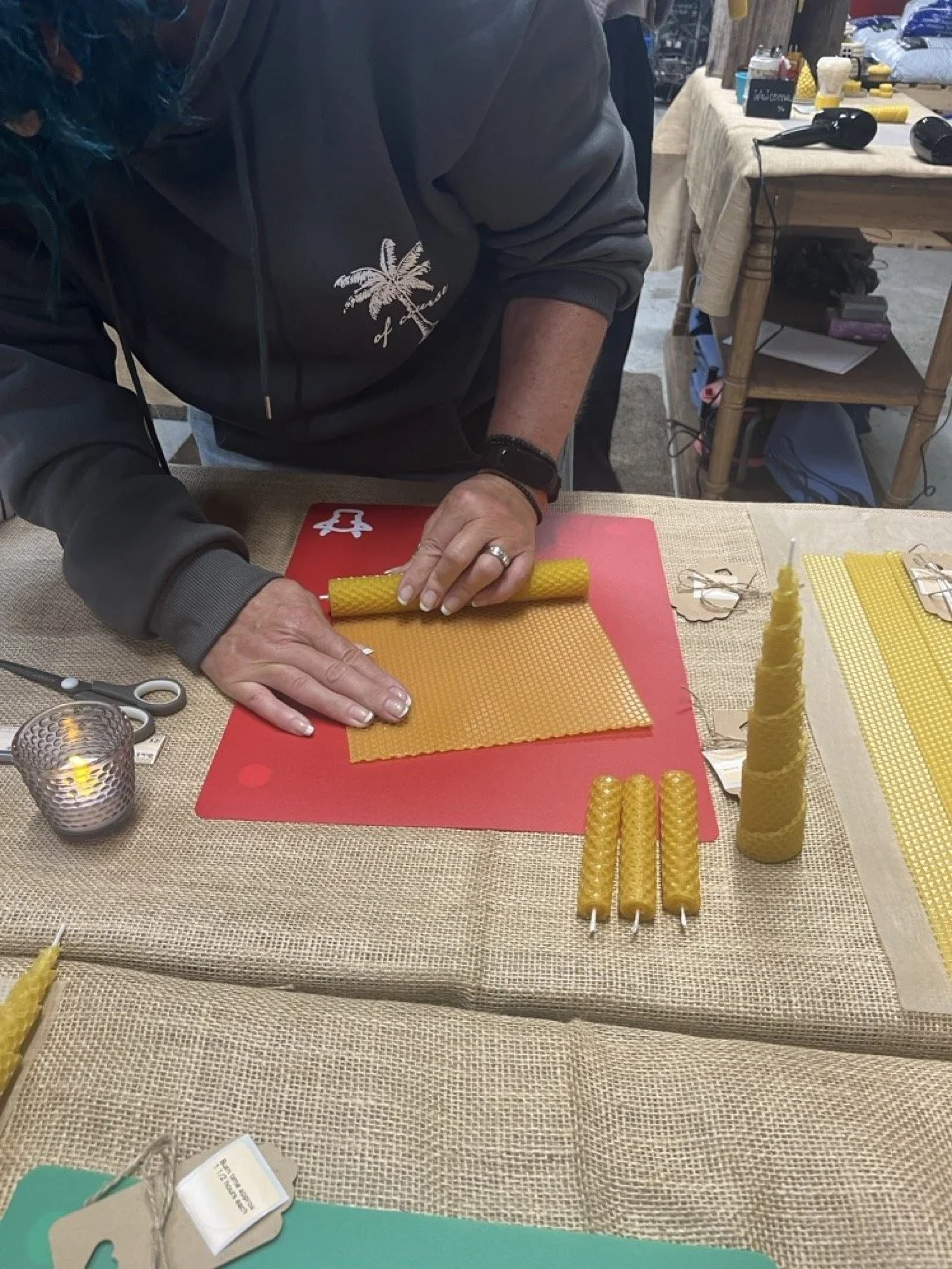 Person rolling yellow beeswax candles on a crafting table with various candles, scissors, and supplies around.