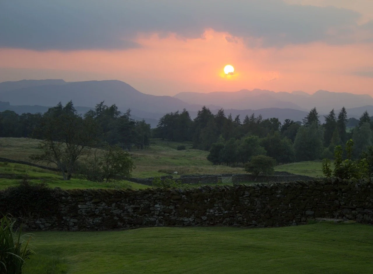Sunset over a landscape with distant mountains, green trees, and a stone wall in the foreground.