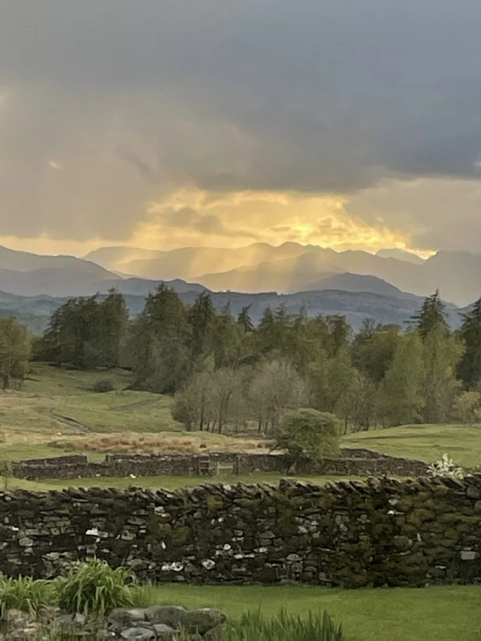 Scenic landscape with rolling green fields, trees, a stone wall, and mountains in the distance under a cloudy sky with rays of sunlight breaking through.