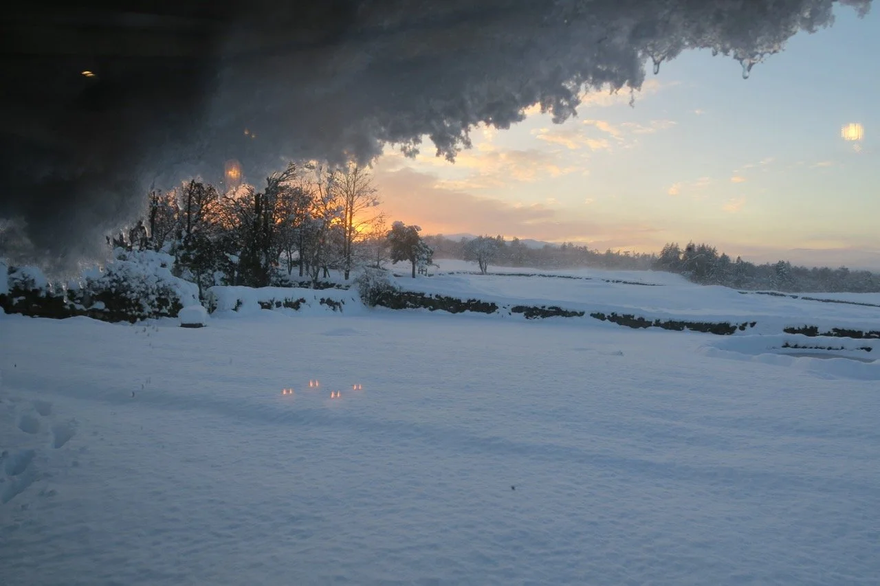 Snow-covered landscape with view of trees and hills during sunset, seen through a window with frost or snow at the top edge.
