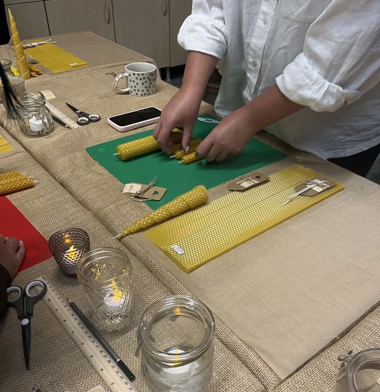 Person cutting yellow beeswax candles on a green cutting mat at a kitchen table, surrounded by crafting supplies, scissors, a smartphone, and lit candles in jars.
