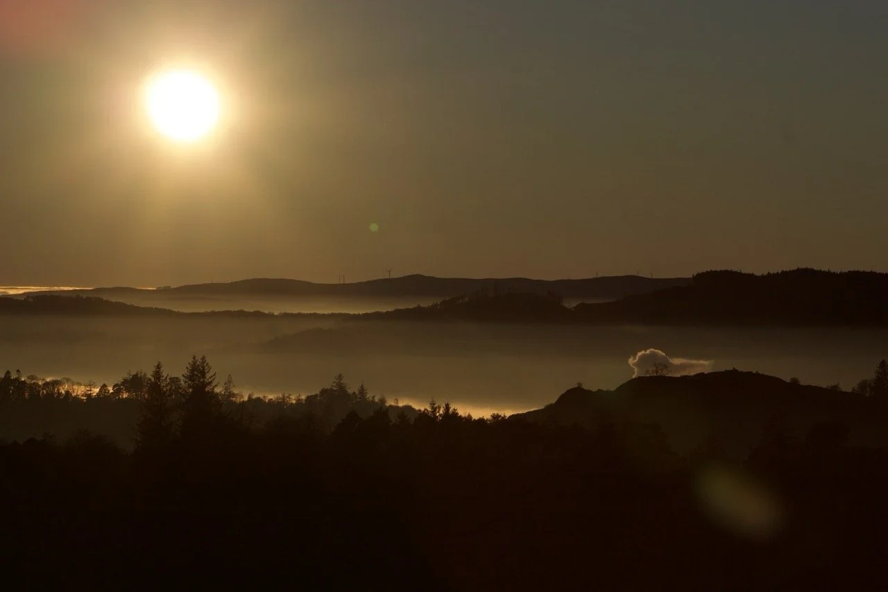Sunset over layered hills with fog, trees in the foreground, and a cloud near the right side.