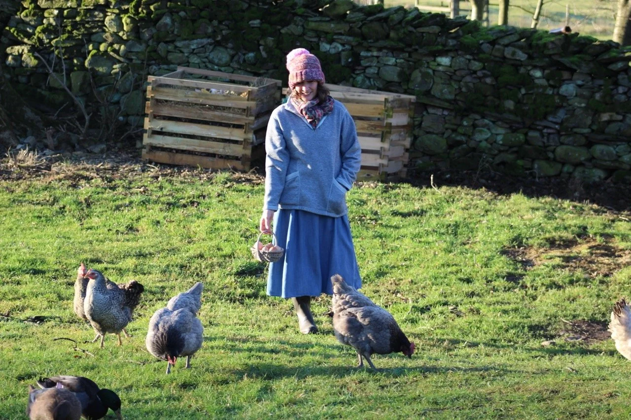 A woman walking in a grassy field with chickens around her, holding a small basket, with stone walls and wooden crates in the background.