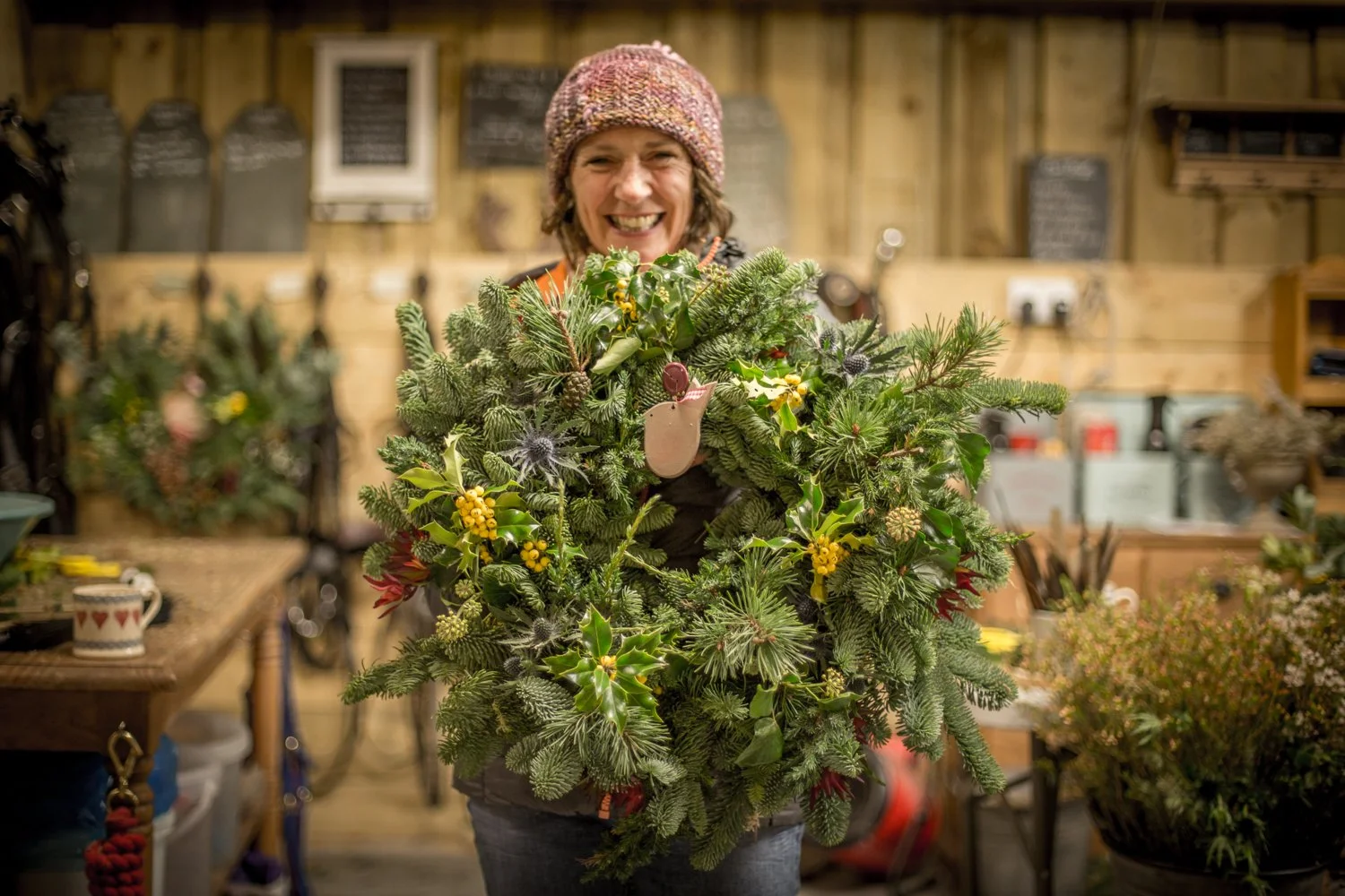 A woman with a colorful knit hat smiling while holding a large festive Christmas wreath in a workshop