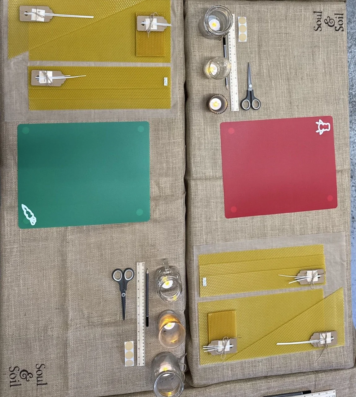 Crafting or quilting setup with colorful sheets, candles, scissors, rulers, and baking supplies on a beige tablecloth.