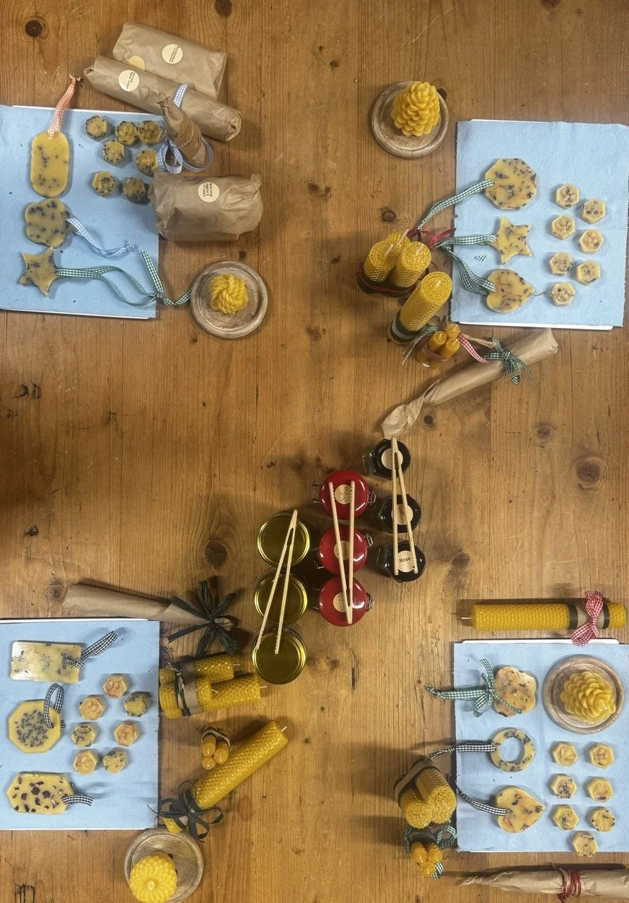 Display of beeswax candles, some wrapped in brown paper, arranged with small honey comb-shaped and star-shaped cookies on blue napkins, and jars of honey on a wooden table.