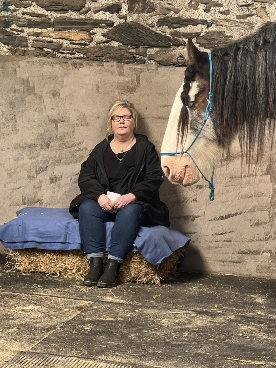 A woman with glasses sitting on a hay bale covered with a blue blanket in a barn, with a large white and brown horse standing nearby.