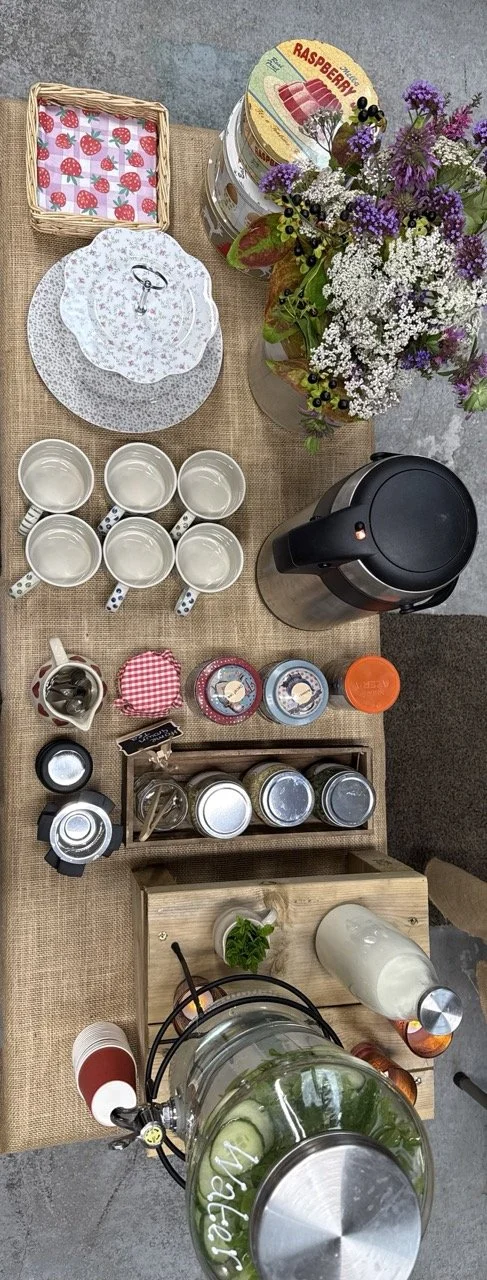 A top-down view of a beverage and snack station on a table with a burlap tablecloth. The station includes a large glass beverage dispenser with sliced cucumbers and a black spigot, six white mugs with polka dot handles, a set of stacked glass jars, a