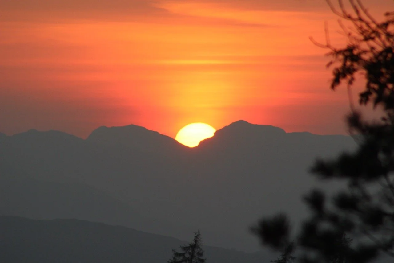 Sunset over mountain range with the sun partially hidden behind the peaks, colorful sky with pink and orange hues, and dark silhouettes of trees in the foreground.