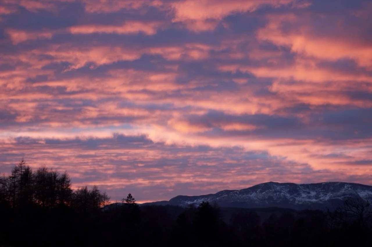 Sunset sky with pink and purple clouds over mountain range and silhouette of trees.