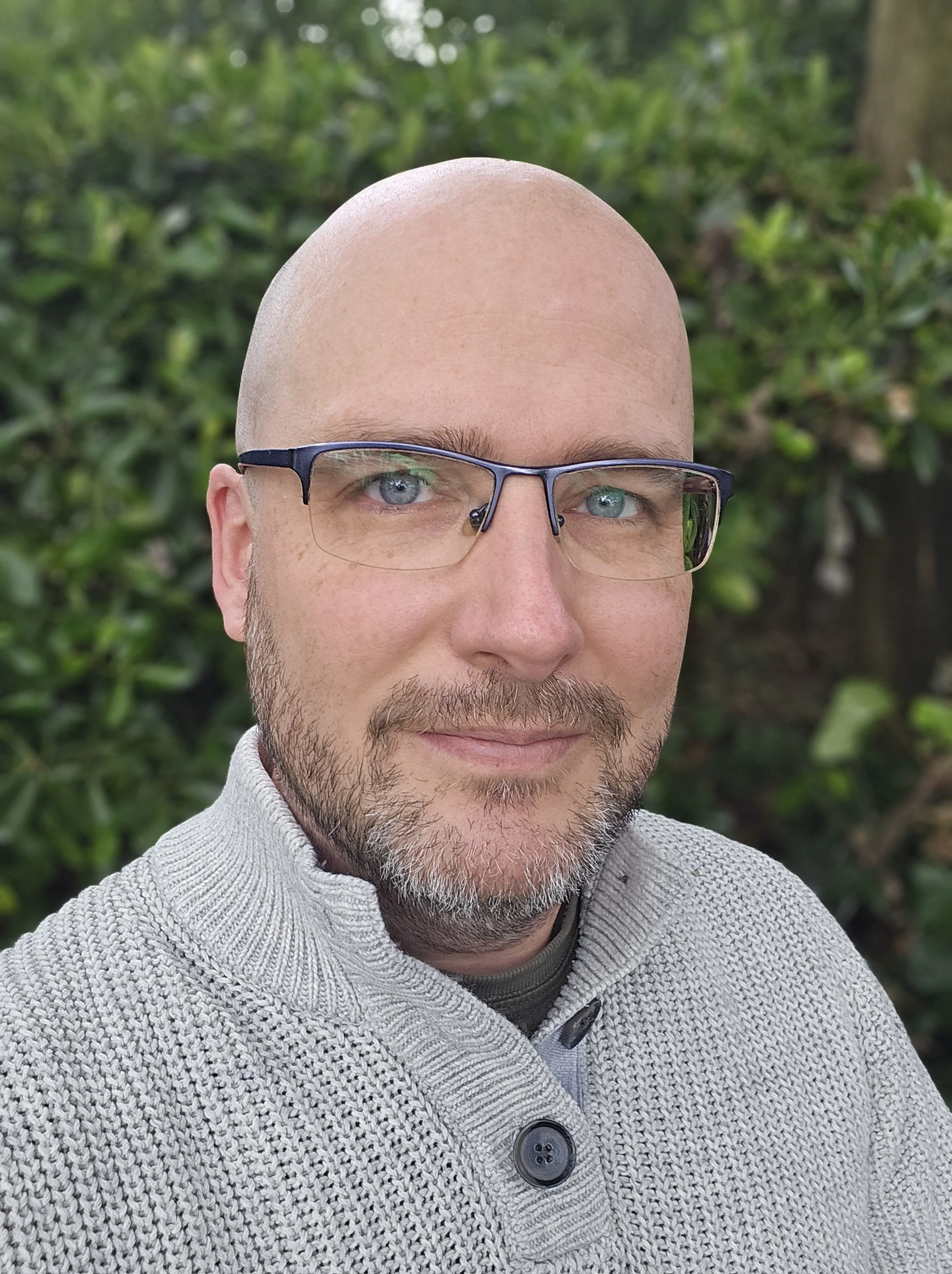A man with a shaved head, light beard, glasses, and blue eyes taking a selfie outdoors, with green foliage in the background. He is wearing a light-colored, textured jacket with large buttons.