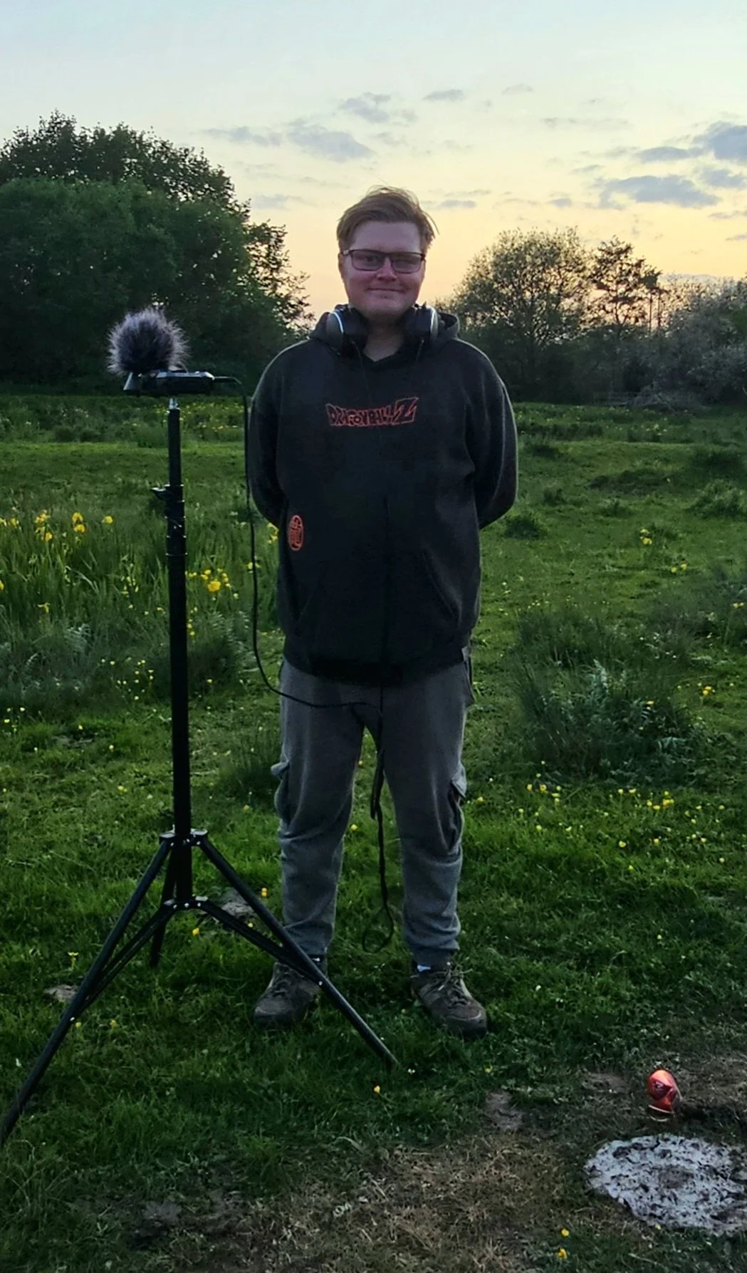 A young man with glasses, smiling, standing outdoors at sunset in a grassy field with trees, wearing a black hoodie and gray pants, with a portable audio recording device on a tripod beside him.