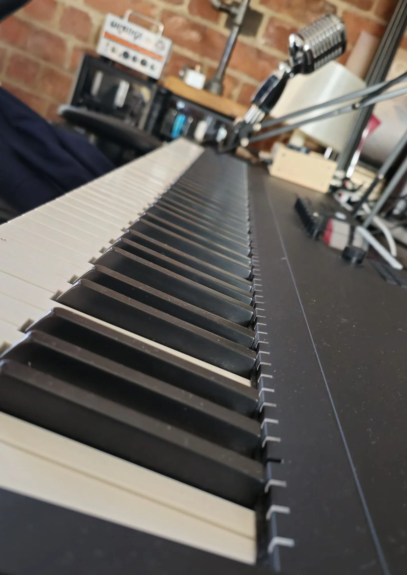Close-up view of a piano keyboard, showing black and white keys, with blurred electronic equipment and a brick wall in the background.