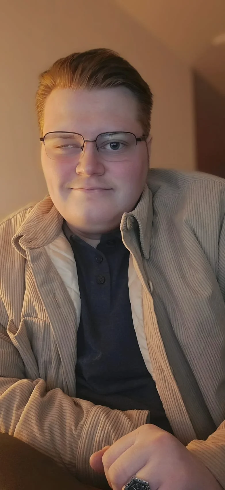 A young man with pale skin, glasses, and short, styled hair sitting indoors. He is smiling slightly, one eye closed, wearing a beige corduroy jacket over a dark shirt, with a ring on his finger.