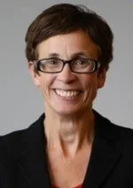 The image is of Penny Pereira. She has short dark hair, glasses and is wearing a black suit