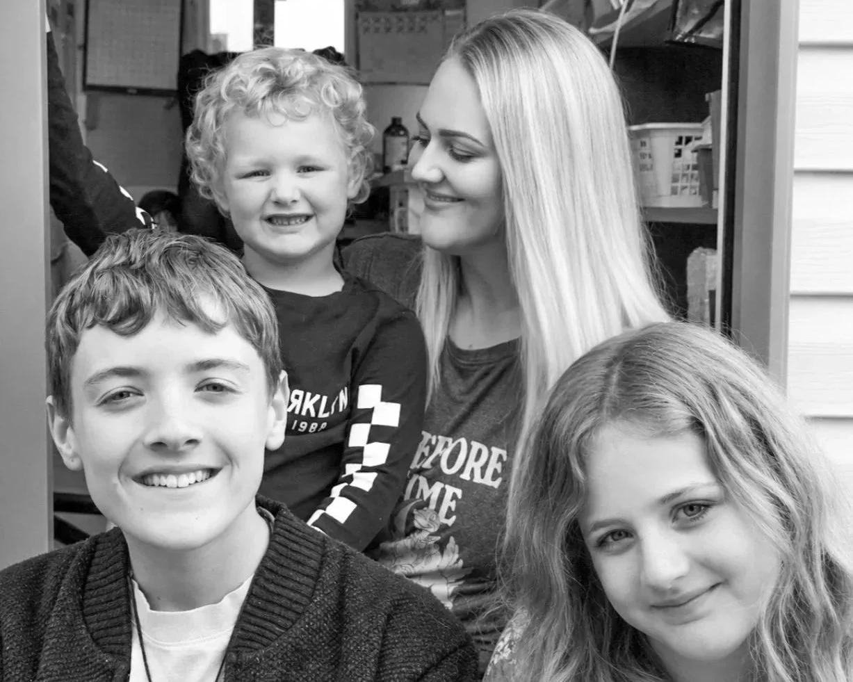 A woman with long blond hair smiling at three children, two boys and a girl, in a group portrait indoors.