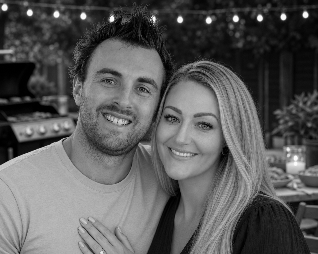 A smiling couple at an outdoor gathering, with a grill and string lights in the background.