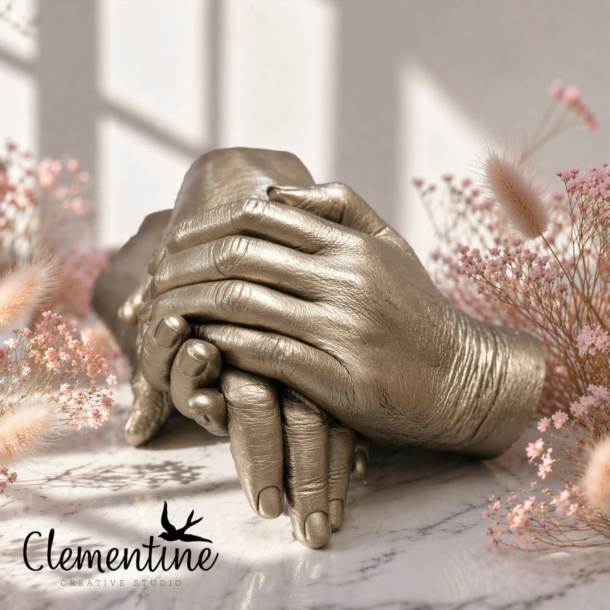 A gold family hand casting featuring adult and baby hands layered together, displayed on marble with pink dried florals — a meaningful bereavement keepsake handcrafted by Clementine Creative Studio, New Zealand.