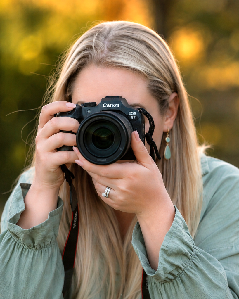 A woman with long blonde hair holding a Canon EOS R7 camera and taking a photo outdoors during sunset. She is wearing a light green jacket and turquoise earrings, with a blurred background of fall foliage.