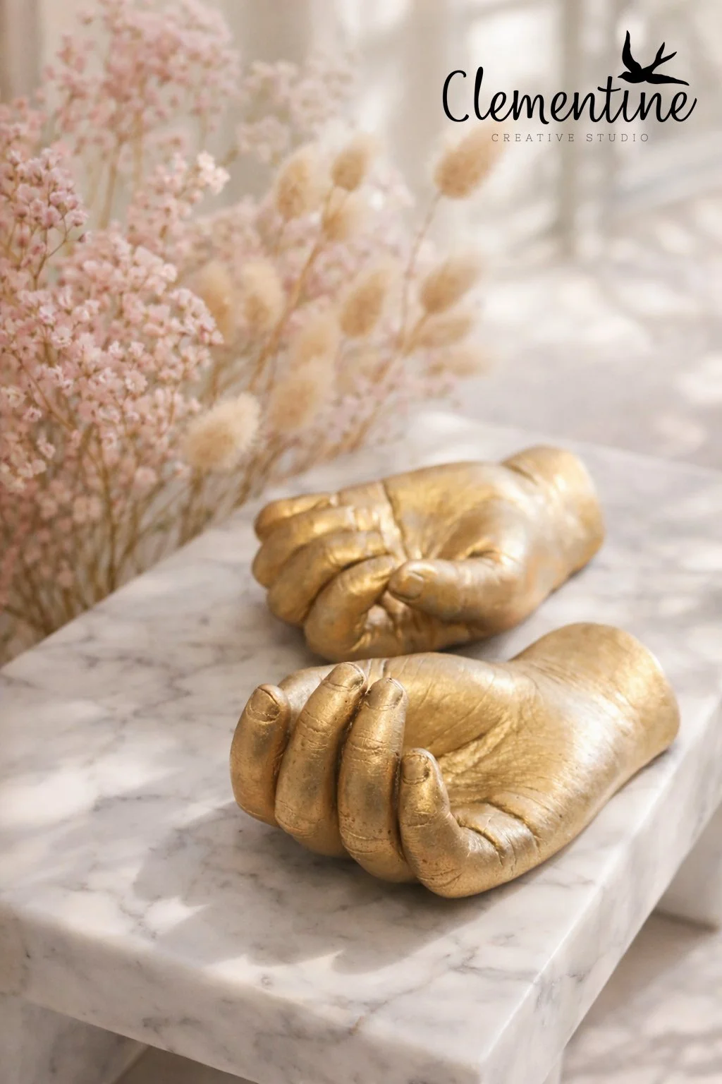 A pair of gold child hand castings resting on a white marble surface with soft pink and beige dried flowers — a tender child bereavement keepsake handcrafted by Clementine Creative Studio, New Zealand.