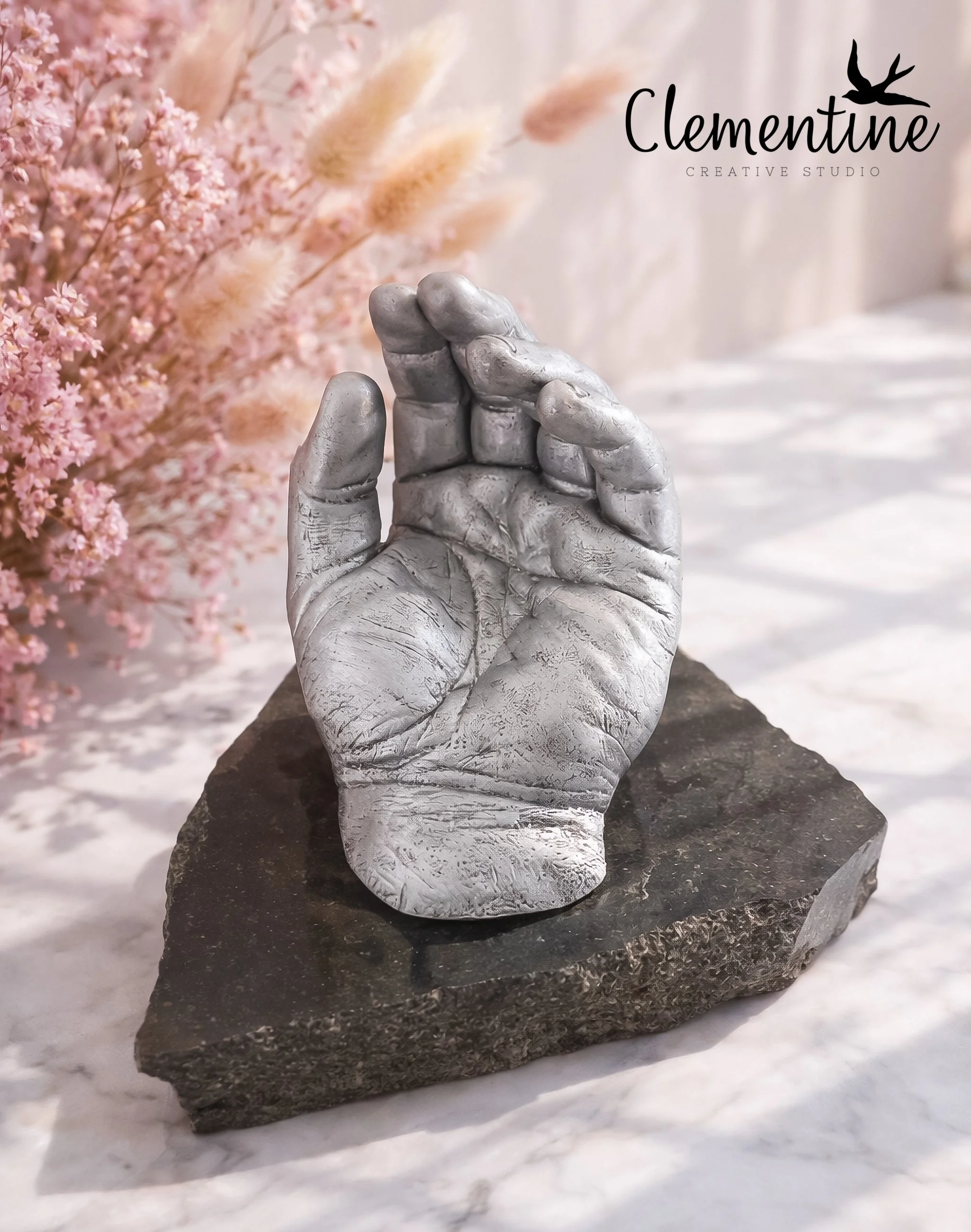 A silver metallic adult hand casting displayed upright on a dark stone base, surrounded by pink dried flowers — a tender bereavement keepsake handcrafted by Clementine Creative Studio, New Zealand.