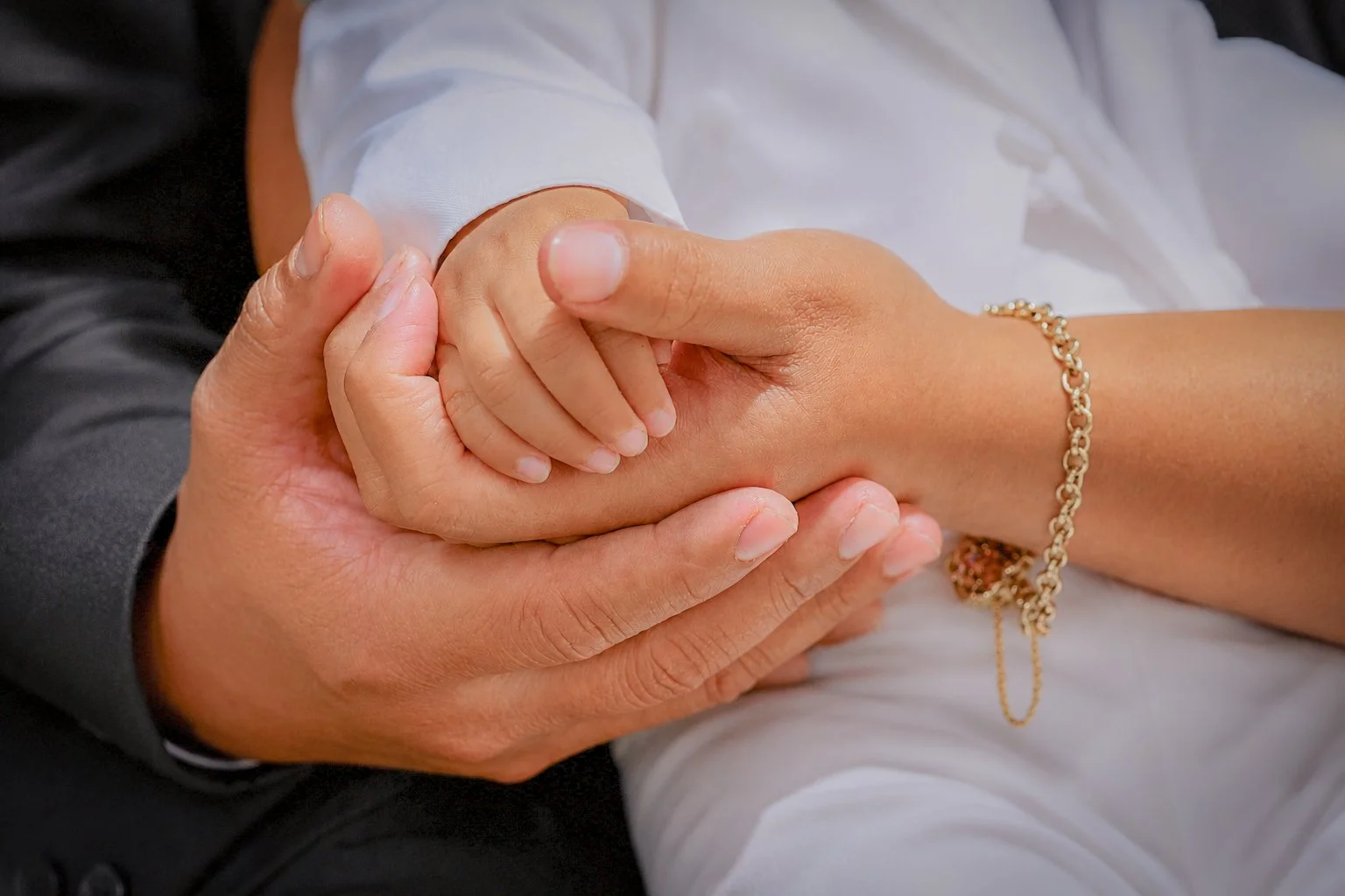 Close-up of a person's hands holding another person's hand for support, with a focus on their intertwined fingers. One person is wearing a white shirt and a gold bracelet, suggesting a warm, supportive gesture.