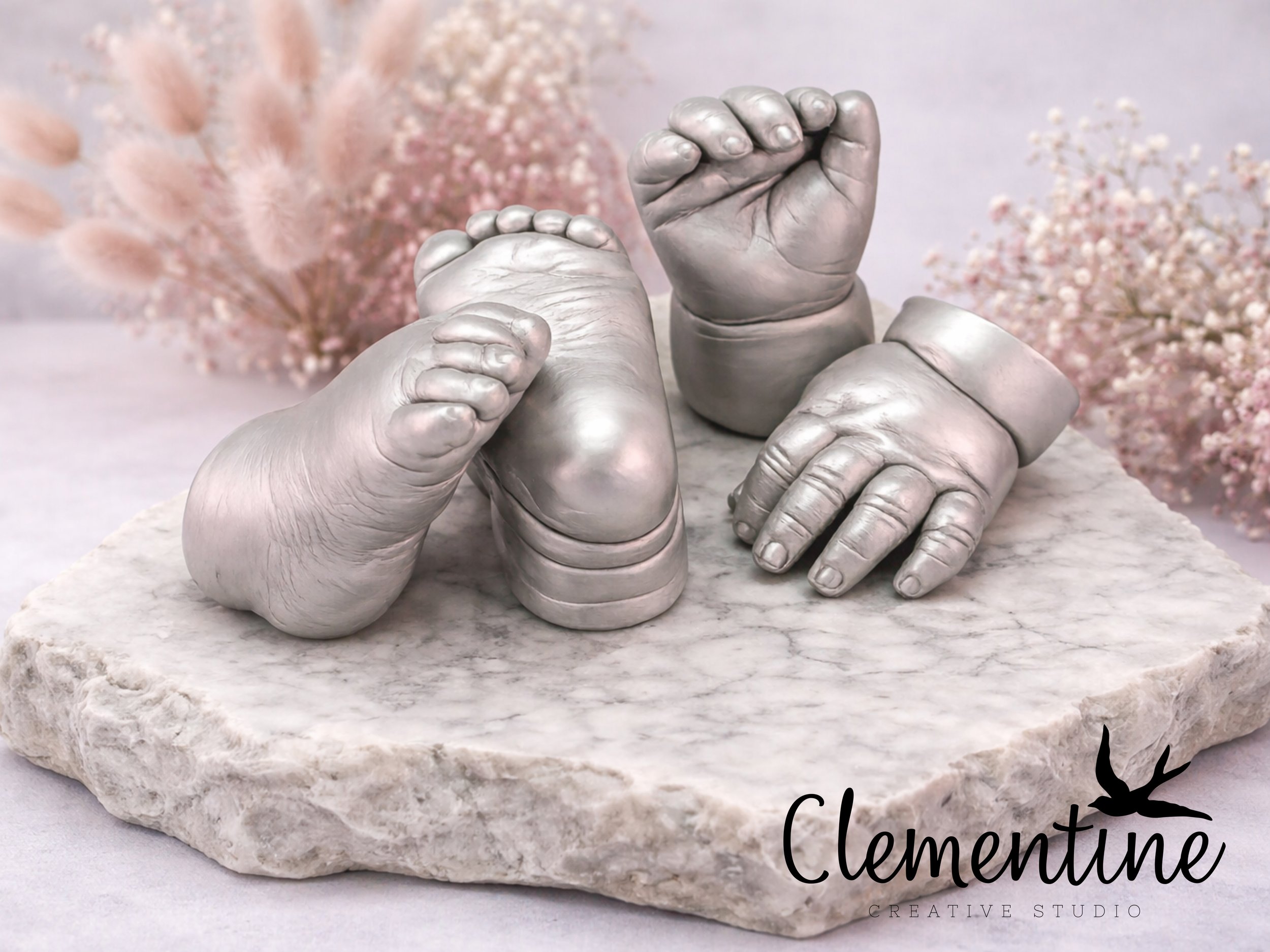 A silver baby hand and foot casting set displayed on a natural marble slab with pink dried flowers — handcrafted by Clementine Creative Studio, New Zealand