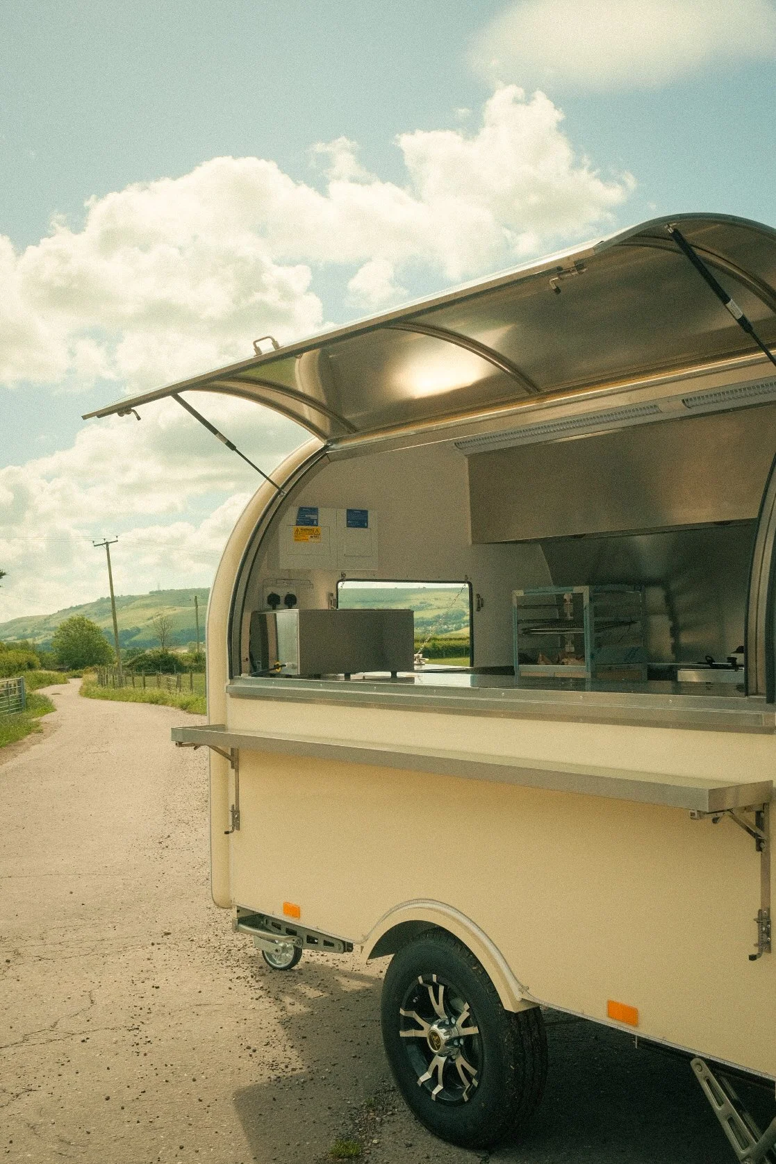 A beige mobile food truck with a large open window and a counter, parked outdoors with a rural landscape, green hills, and a cloudy sky in the background.