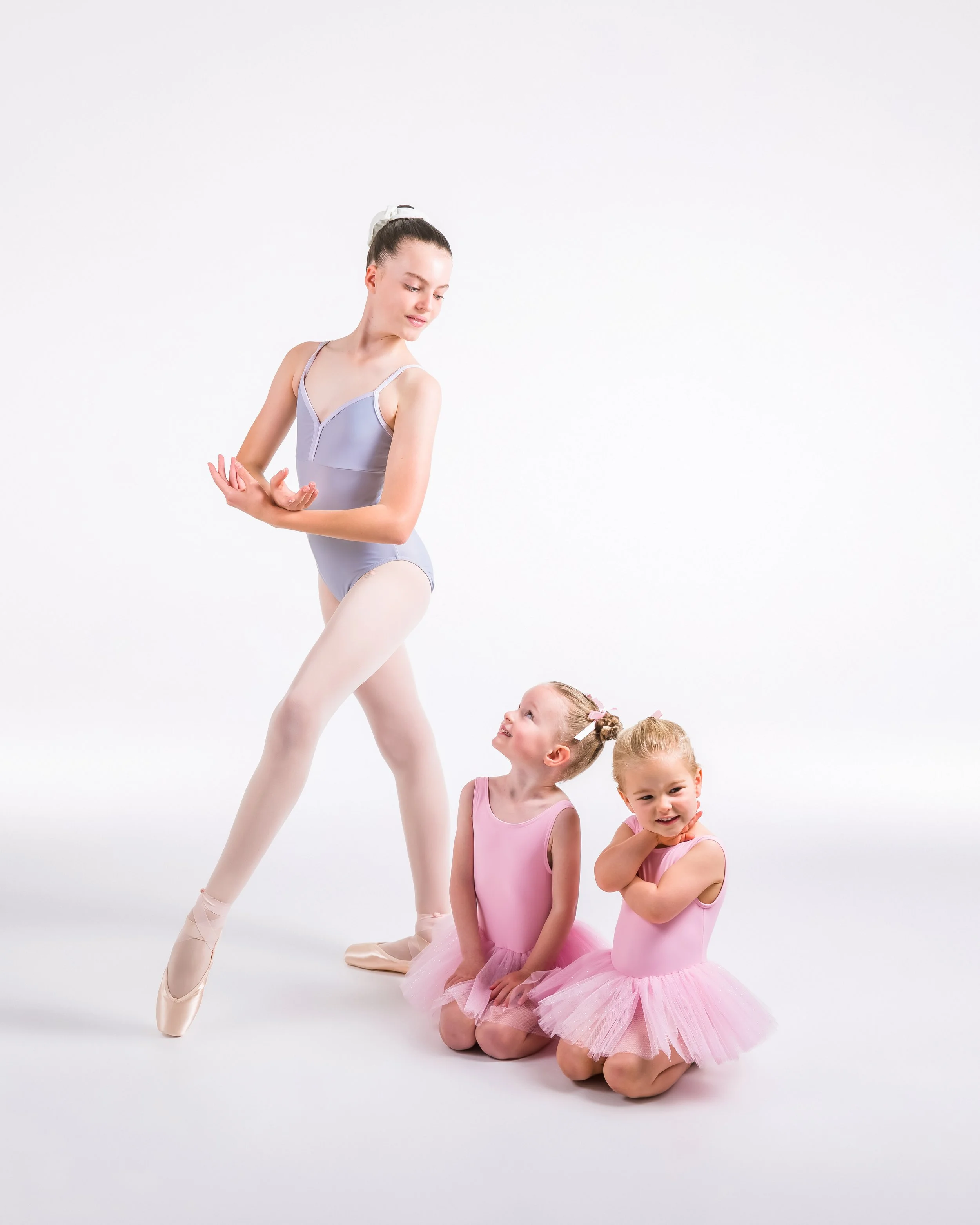 A ballet studio scene with three young girls in pink tutus kneeling on the floor, and an older girl in ballet attire standing with one leg slightly bent and her arms crossed behind her back, all against a plain white background.