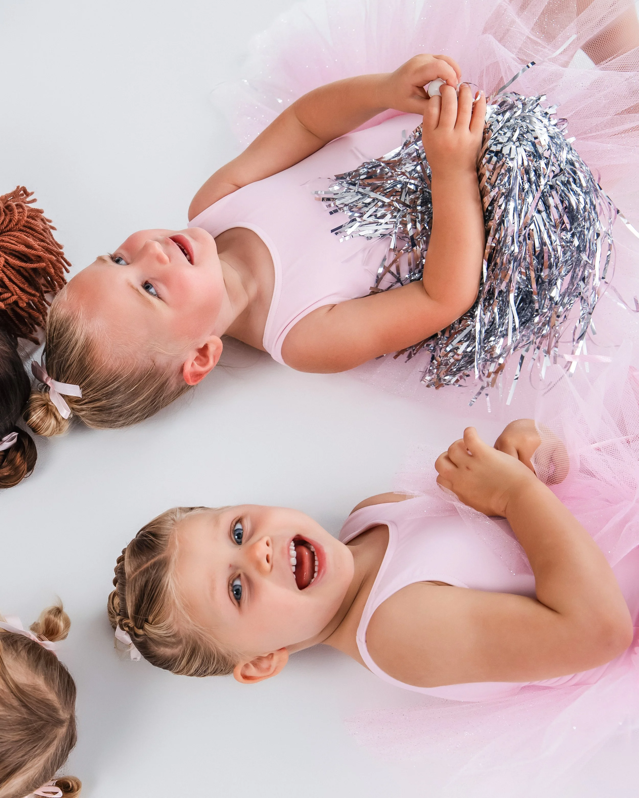 A group of young girls with blonde hair styled in braids, wearing pink ballet tutus, lying on a white surface, smiling and holding metallic silver and blue pom-poms.