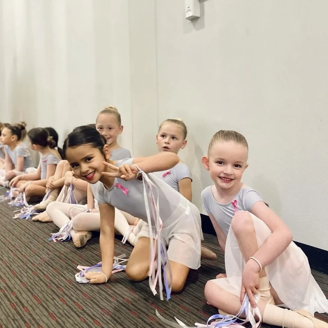 Group of young girls sitting on the floor in ballet costumes, smiling and making peace signs.