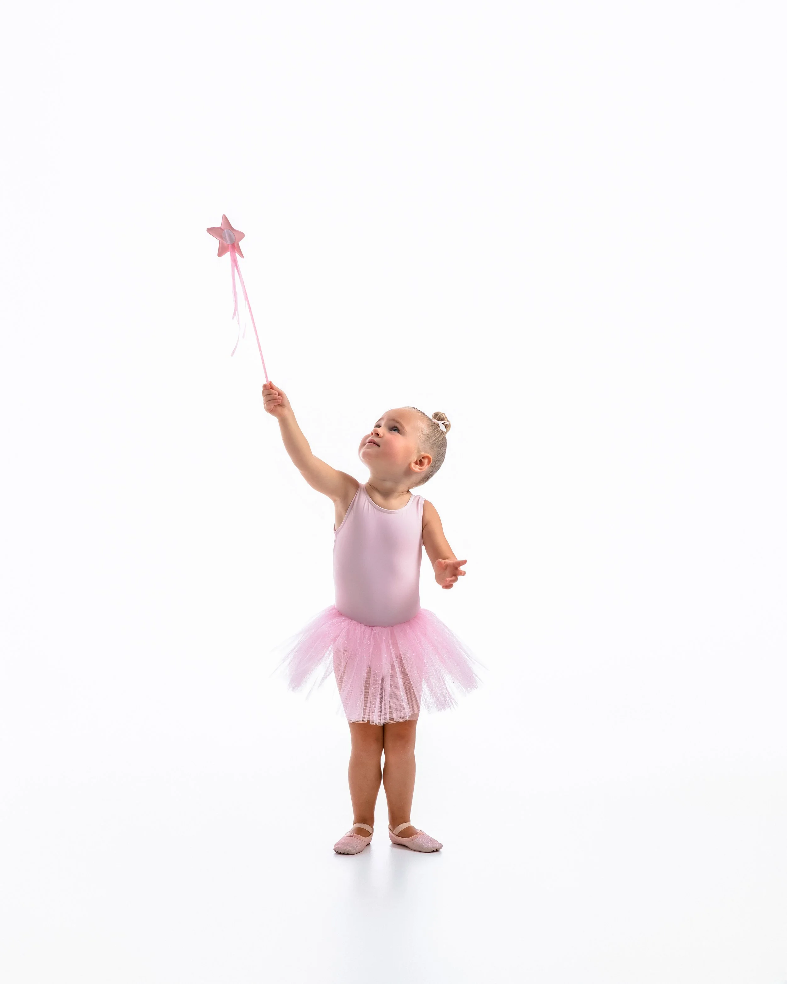 Young girl in a pink ballet tutu and ballet slippers, holding a pink star-shaped wand, standing against a plain white background.