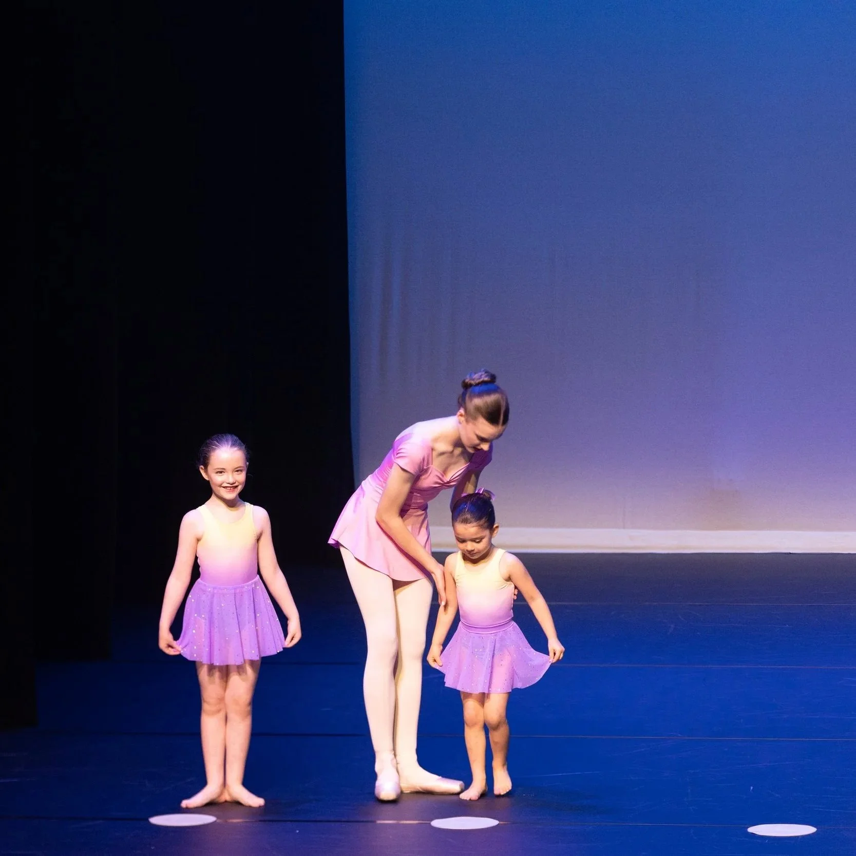 Ballet class with four young girls in purple dresses and a woman in a pink ballet outfit on stage with a blue background.