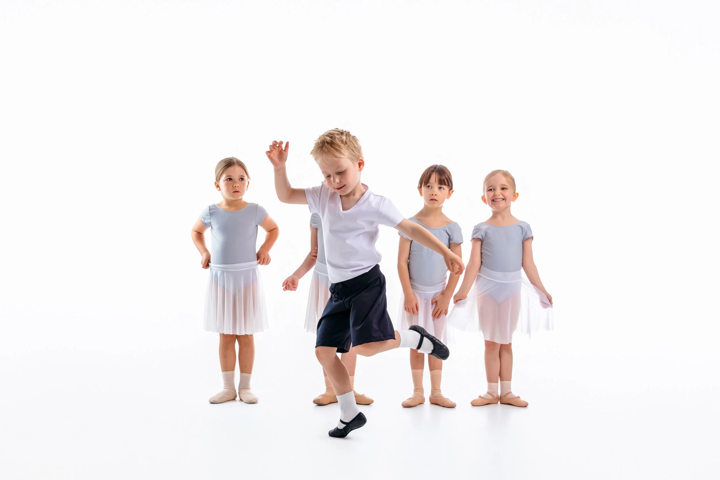 Four young girls and one boy in ballet attire, with the boy in the center leaping or dancing while two girls watch and two others smile, all against a plain white background.