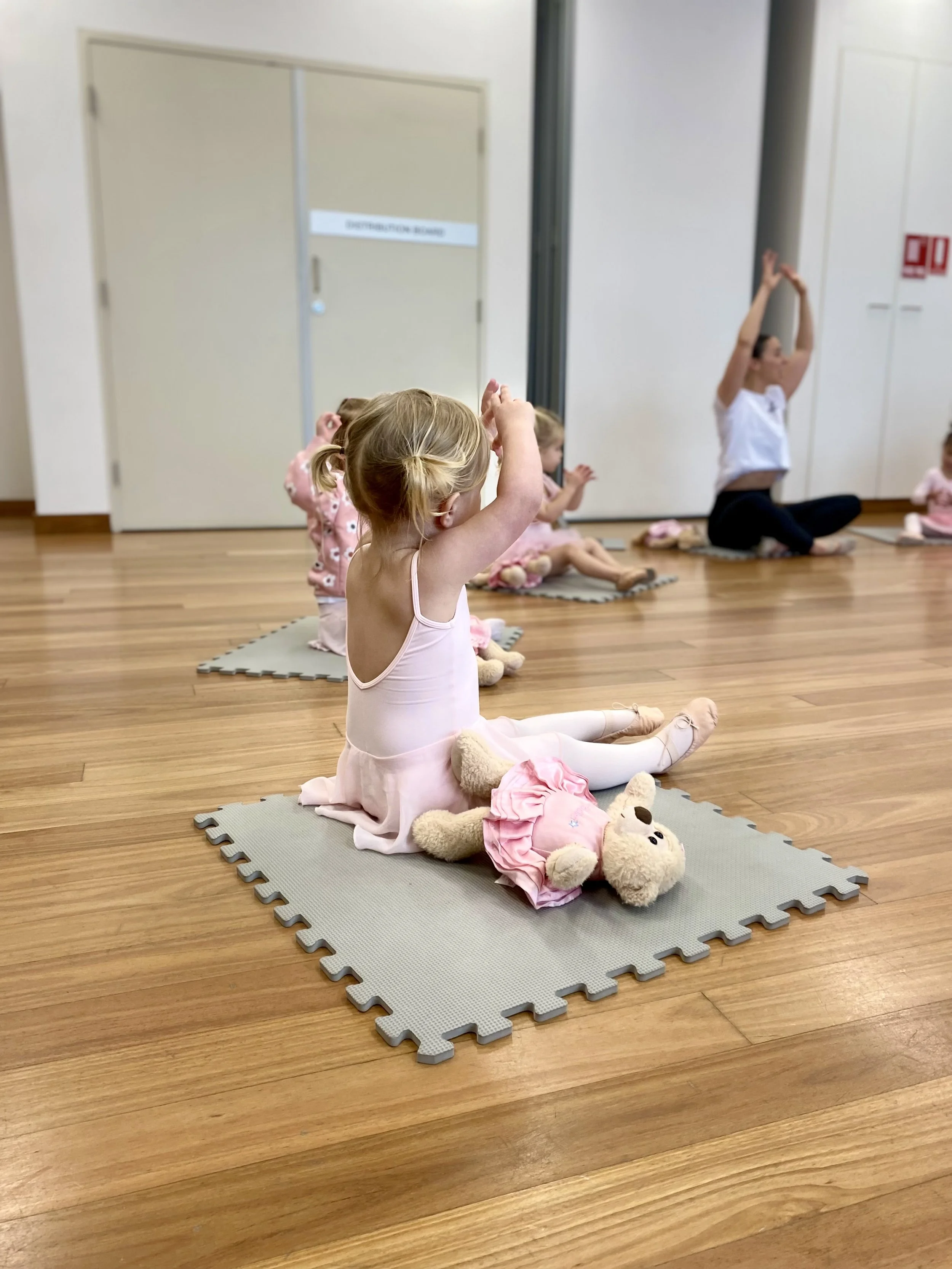 Children in a ballet class sitting on mats with stuffed animals, practicing ballet moves with an instructor in a studio with wooden floors.