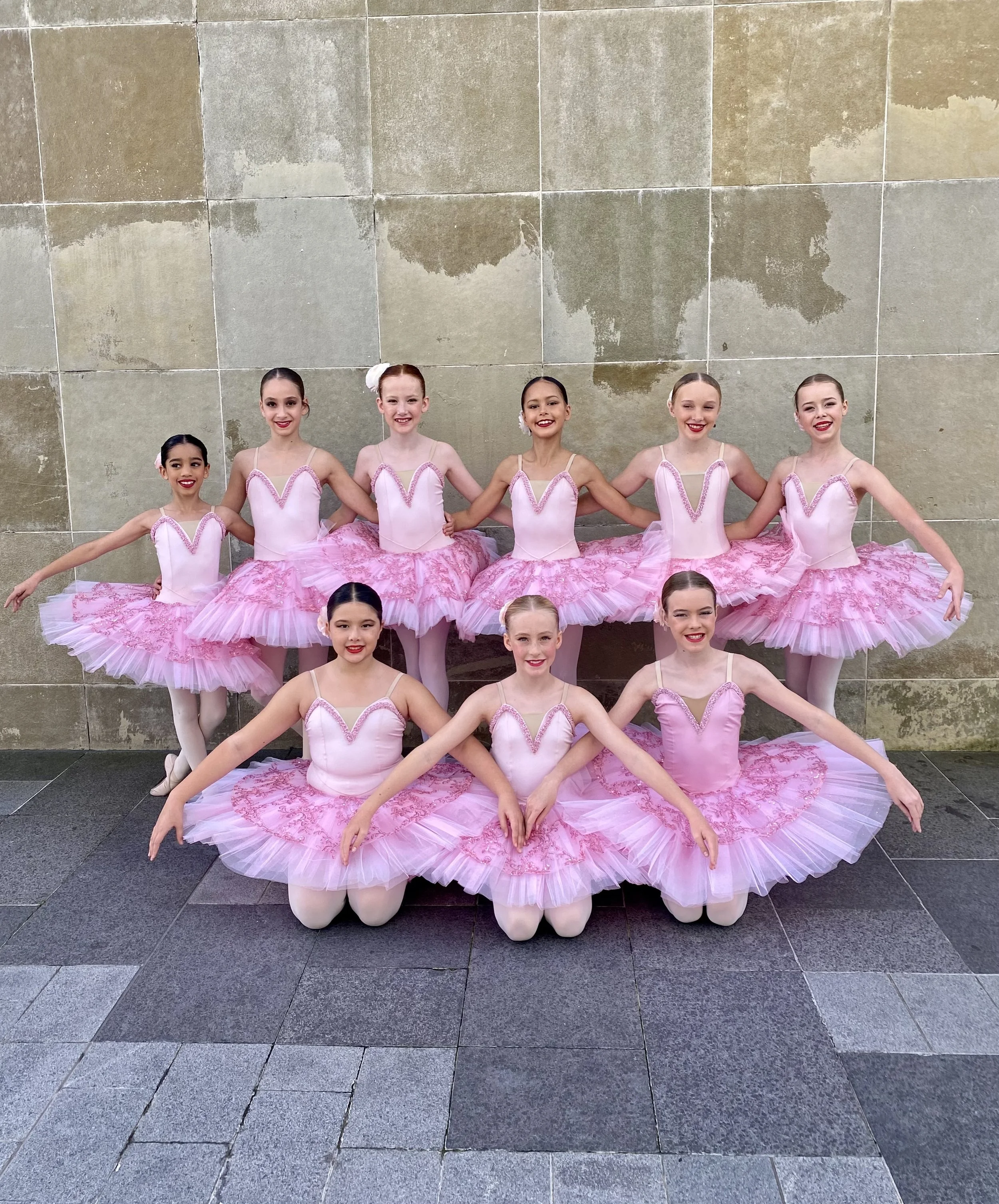 Group of young ballet dancers in pink tutus and leotards, kneeling with arms around each other, posed against a beige stone wall.