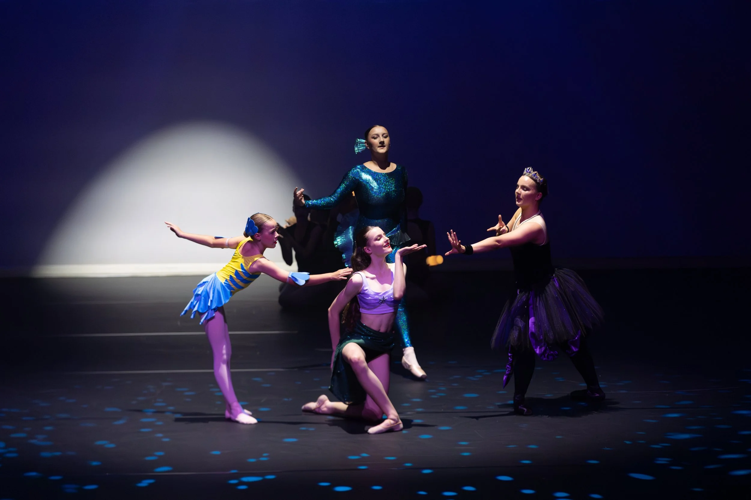 A group of five ballet dancers performing on a dark stage. One dancer is standing in the center, wearing a shimmering blue costume and a crown. The other four dancers are crouched or kneeling around her, dressed in colorful costumes, with one in a yellow and blue outfit, another in a pink top with black shorts, and two in black costumes. The dancers are engaged in a coordinated performance with expressive gestures and poses.
