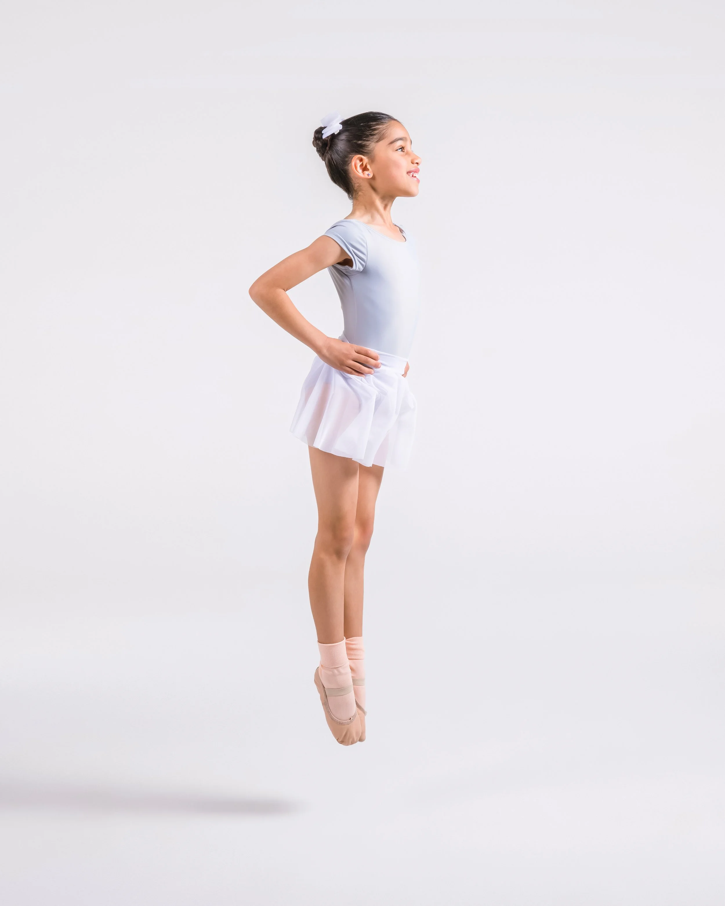 Young girl dressed as a ballet dancer standing on pointe with her hands on her hips, facing to the right, against a plain white background.